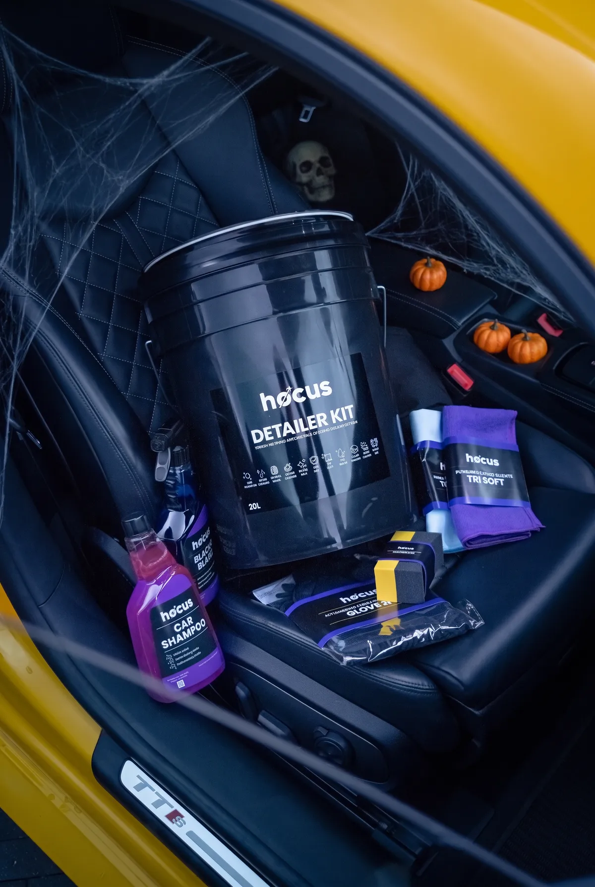 HOCUS Detailer Kit with car care bottles, applicator and microfibre cloths on a yellow car’s black leather seat, with Halloween pumpkins and spiderwebs in the background